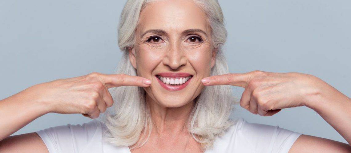 avoid-these Older woman with long gray hair smiling and pointing at her teeth with both index fingers, wearing a white shirt and standing against a plain light background, showcasing her bright smile after visiting dental Grande Prairie.