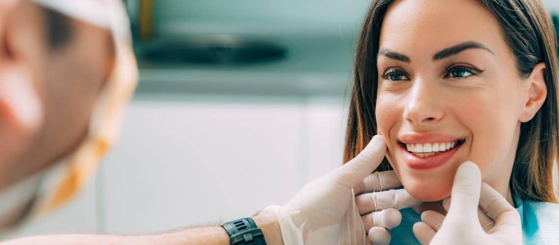 cosmetic-dentist A dentist at a dental grande prairie clinic, wearing gloves, examines a smiling woman's teeth during a dental check-up.