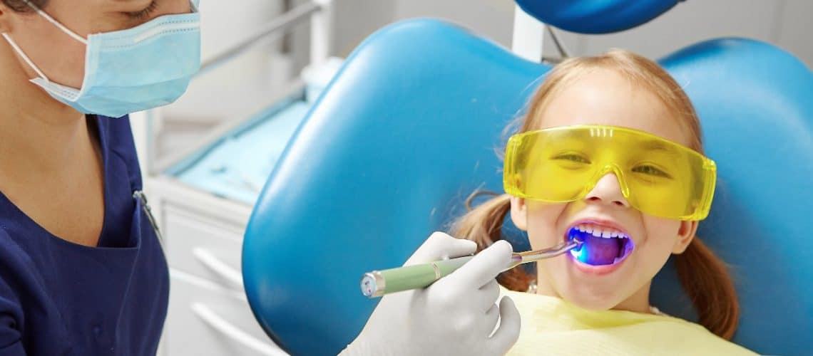 A dentist at Dental Grande Prairie uses a curing light on a young girl wearing yellow protective glasses while she sits comfortably in the dental chair.
