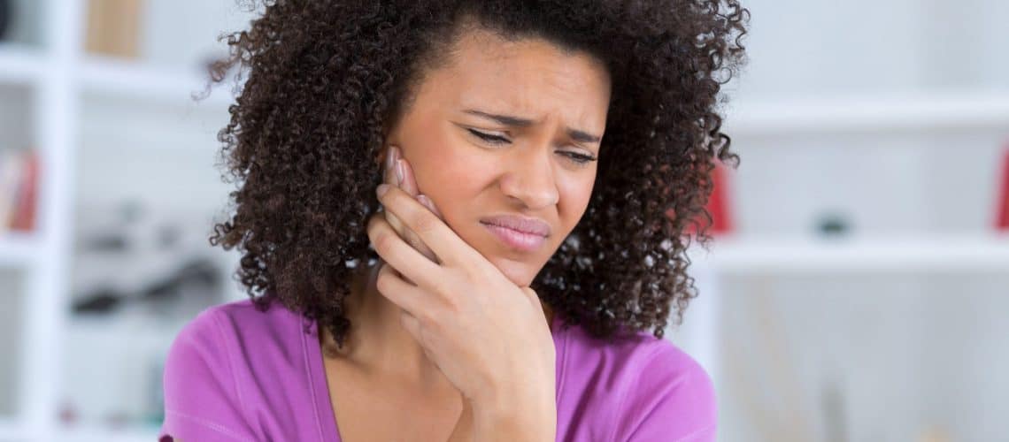 emergencies A person with curly hair wearing a purple top holds their cheek and appears to be in pain, possibly experiencing a toothache and in need of dental Grande Prairie services.