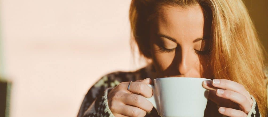 how-can-i-remove-coffee-stains-from-my-teeth A woman with long blonde hair holds a white mug with both hands and drinks from it, wearing a patterned sweater in soft natural light—her bright smile hinting at recent dental Grande Prairie care.