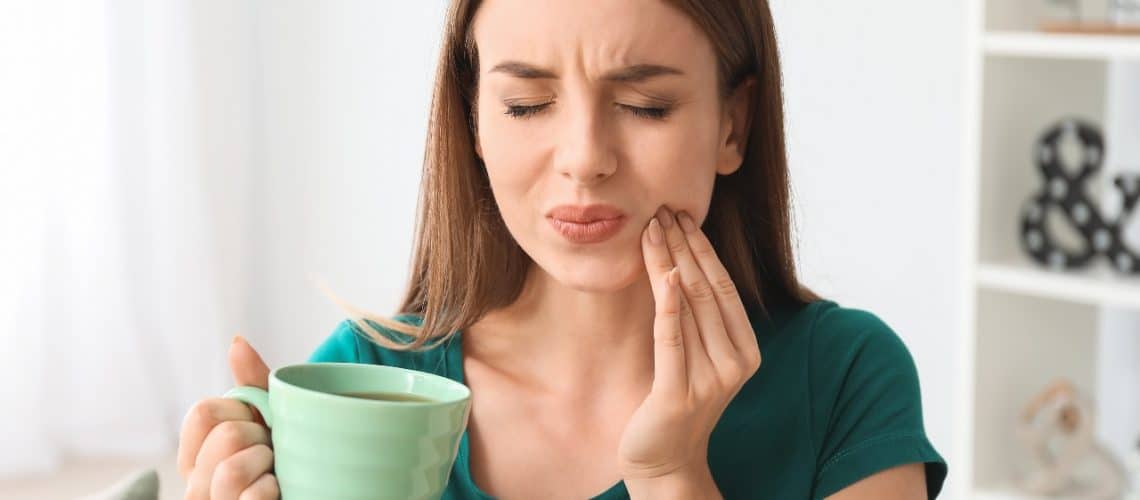 is-tooth-sensitivity-permanently-curable Woman holding a green mug and touching her cheek, appearing to experience tooth or mouth pain—possibly in need of dental care in Grande Prairie.