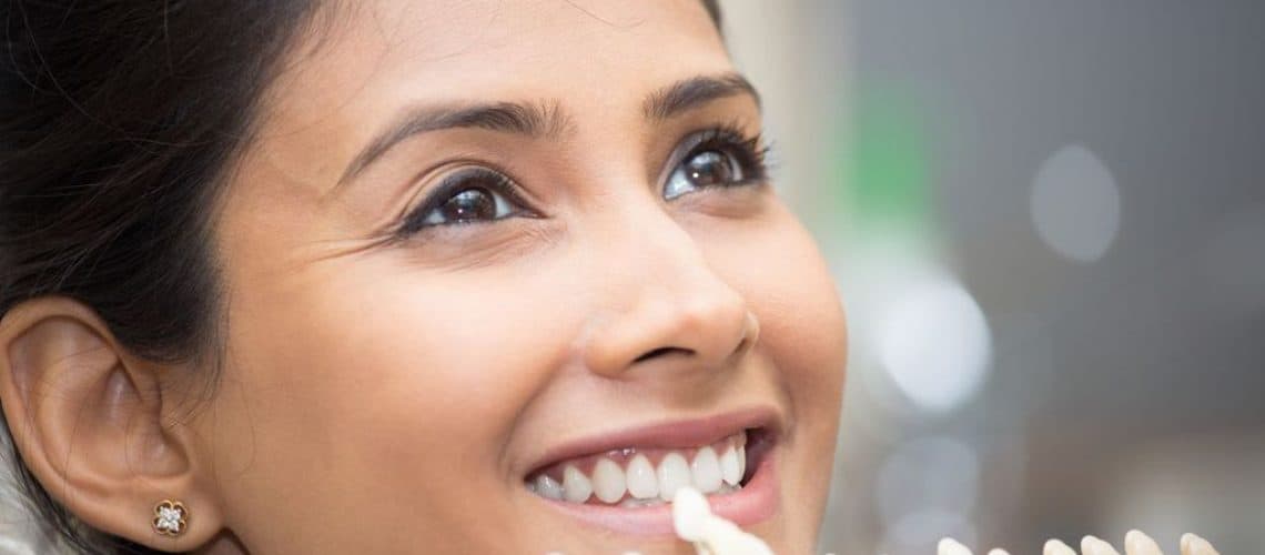 long-do-dental Woman smiling while holding a dental shade guide next to her teeth for color comparison at a dental Grande Prairie clinic.