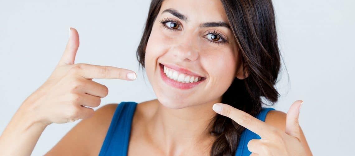 mouth-washes Woman in a blue top smiling and pointing to her teeth with both hands against a plain background, showcasing her bright smile after visiting dental Grande Prairie.
