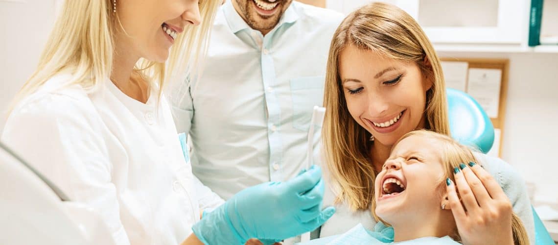 A dentist at a dental Grande Prairie clinic shows a toothbrush to a smiling child sitting on a woman's lap, while an adult man stands nearby.