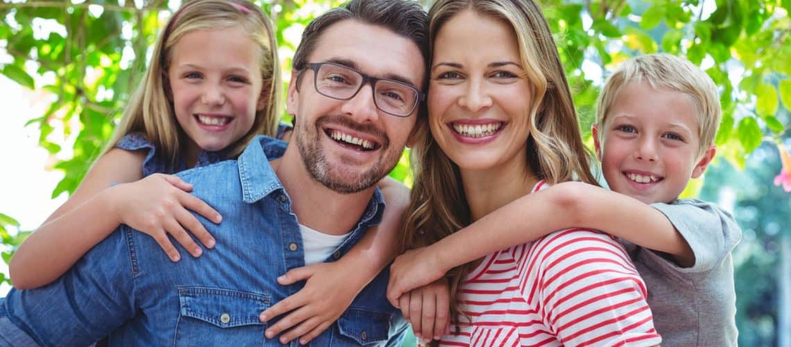 rest-of-the-body Smiling family of four posing outdoors, with two children hugging their parents from behind, surrounded by green foliage—showcasing the joy that comes with healthy smiles from dental Grande Prairie care.