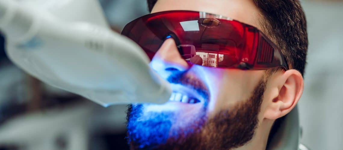side-effects A man wearing protective red glasses receives a teeth whitening treatment with a blue light device at a dental clinic in Dental Grande Prairie.