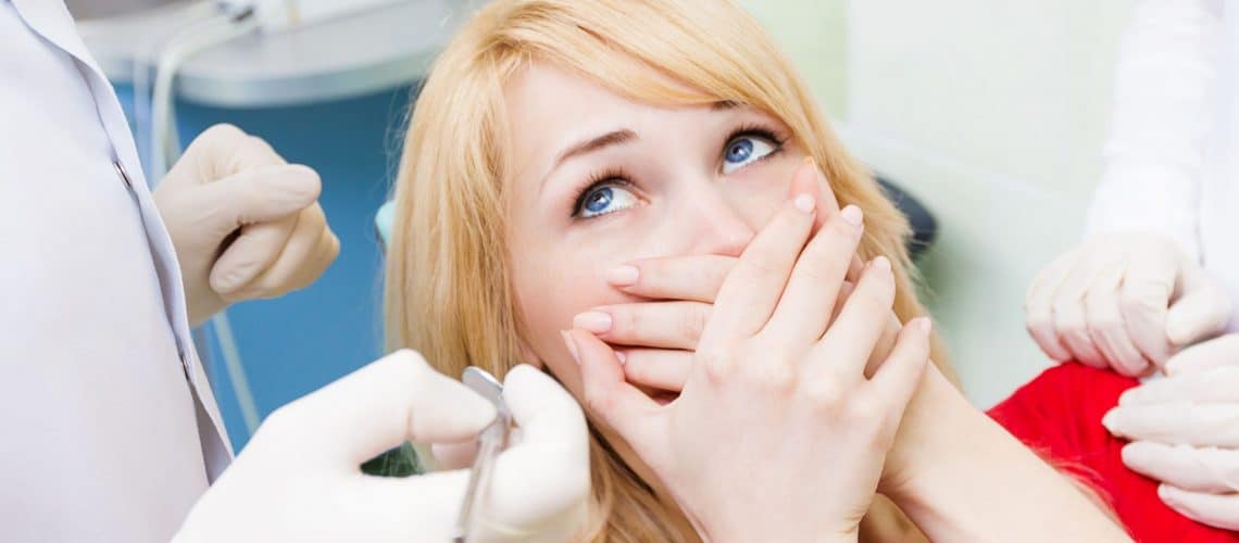 tooth-pain A woman with blonde hair covers her mouth with both hands while sitting in a dental chair at a dental Grande Prairie clinic, looking anxious as a dentist holds dental tools nearby.