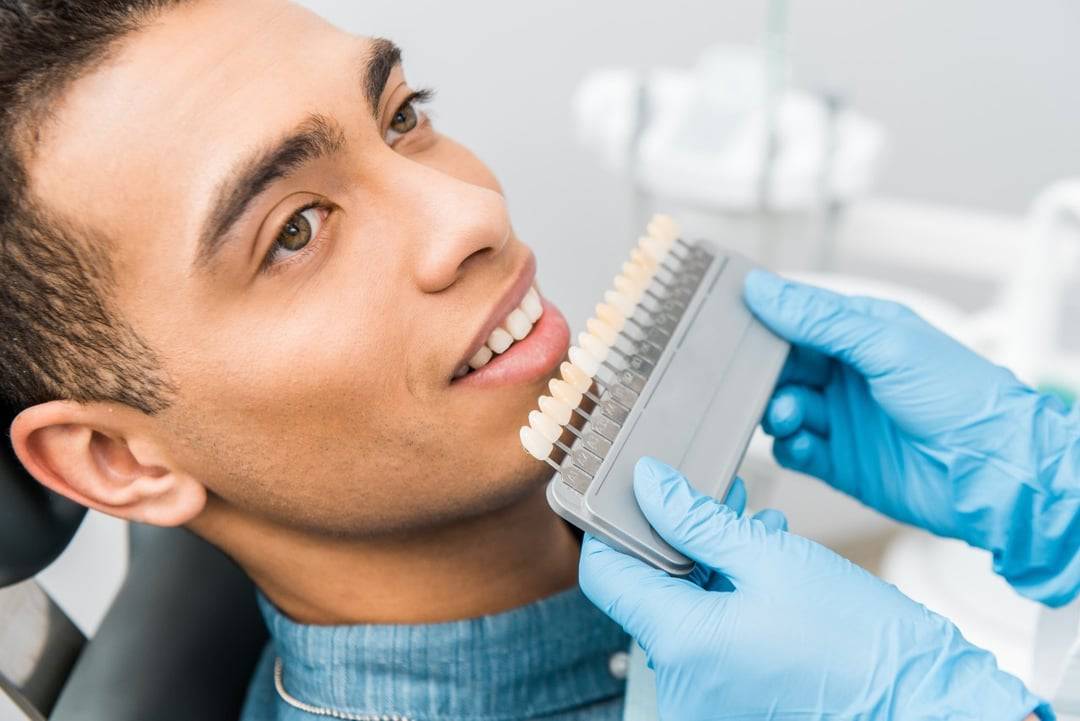 are-porcelain-veneers-better-than-composite-veneers A dentist wearing blue gloves holds a tooth shade guide next to a smiling patient’s teeth at a dental grande prairie clinic.