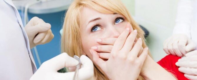 A woman with blonde hair sits in a dental chair, covering her mouth with both hands while two dentists in gloves hold dental instruments nearby at a dental grande prairie clinic.