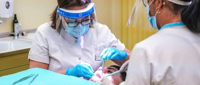 Two dental professionals wearing face shields and masks perform a dental procedure on a patient at a dental Grande Prairie clinic. Dental tools and equipment are visible in the foreground.