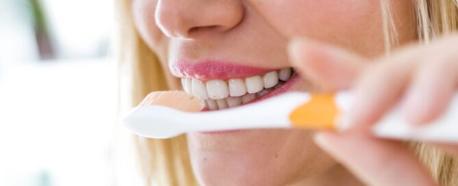 A woman with blonde hair and pink lipstick brushes her teeth with a white and orange toothbrush, maintaining her bright smile just like after a visit to Dental Grande Prairie.