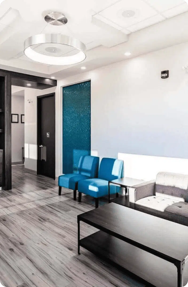 Modern waiting room at a dental Grande Prairie clinic with blue chairs, a gray sofa, sleek black coffee table, wood flooring, and white walls featuring a textured blue accent panel.