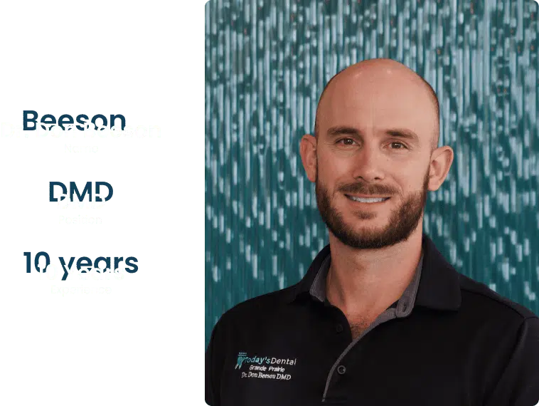 A man with a beard and a shaved head smiles at the camera, standing in front of a blue patterned background. He is wearing a black collared shirt with embroidery, sharing his warm smile at Dental Grande Prairie.