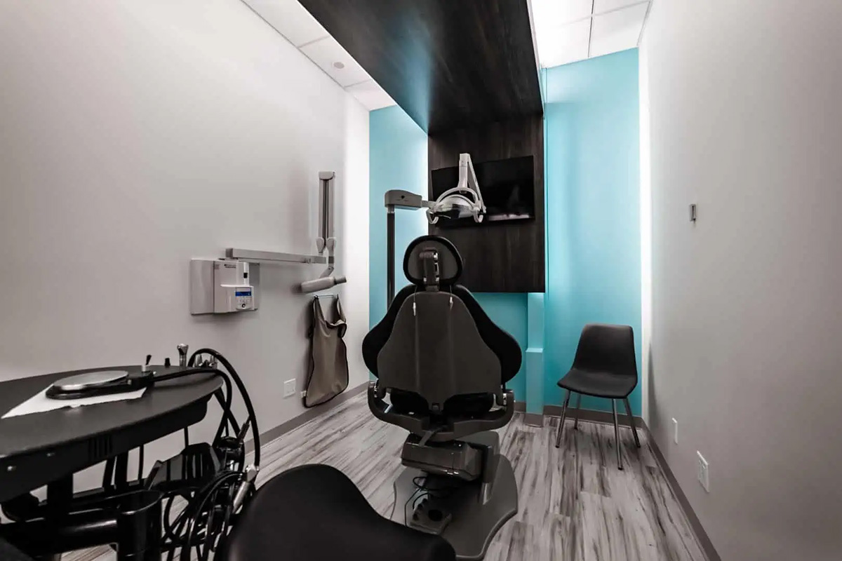 A modern dental exam room in Dental Grande Prairie, featuring a dental chair, equipment, wall-mounted monitor, two gray chairs, and a blue accent wall.