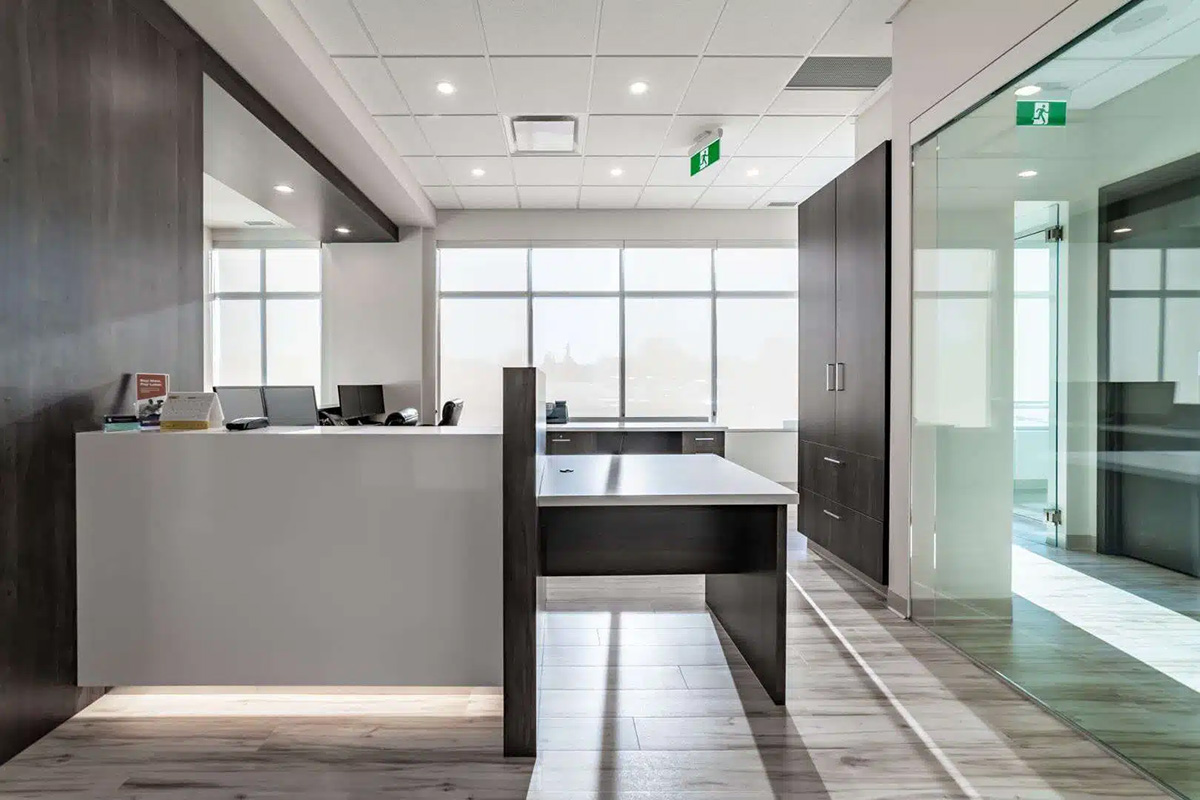 Modern office reception area for a dental Grande Prairie clinic, featuring a front desk, computers, glass walls, and natural light streaming through large windows.