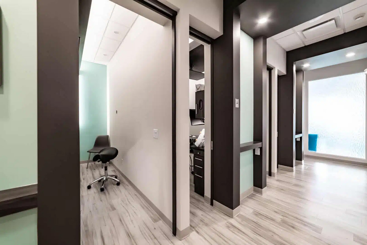 A modern dental Grande Prairie clinic hallway with exam rooms, a rolling stool, light wood flooring, and mint green and white walls.