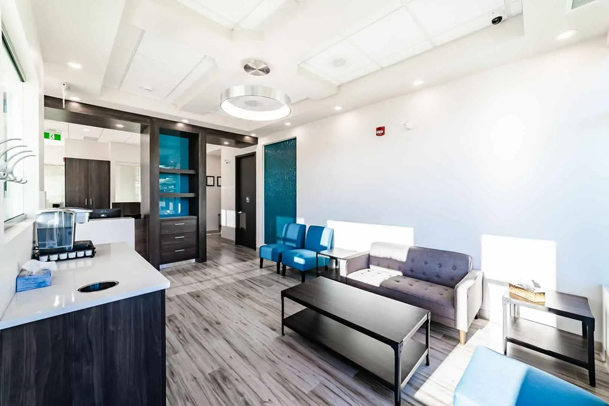 Bright, modern waiting room at Dental Grande Prairie with gray and blue chairs, a coffee station on the left, wood flooring, and large windows letting in natural light.