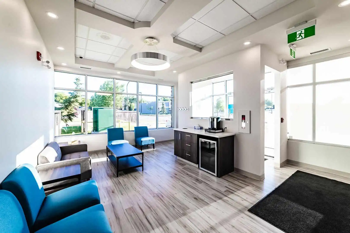 A modern waiting room at Dental Grande Prairie with blue and gray chairs, a coffee station, large windows, wood flooring, and bright overhead lighting.