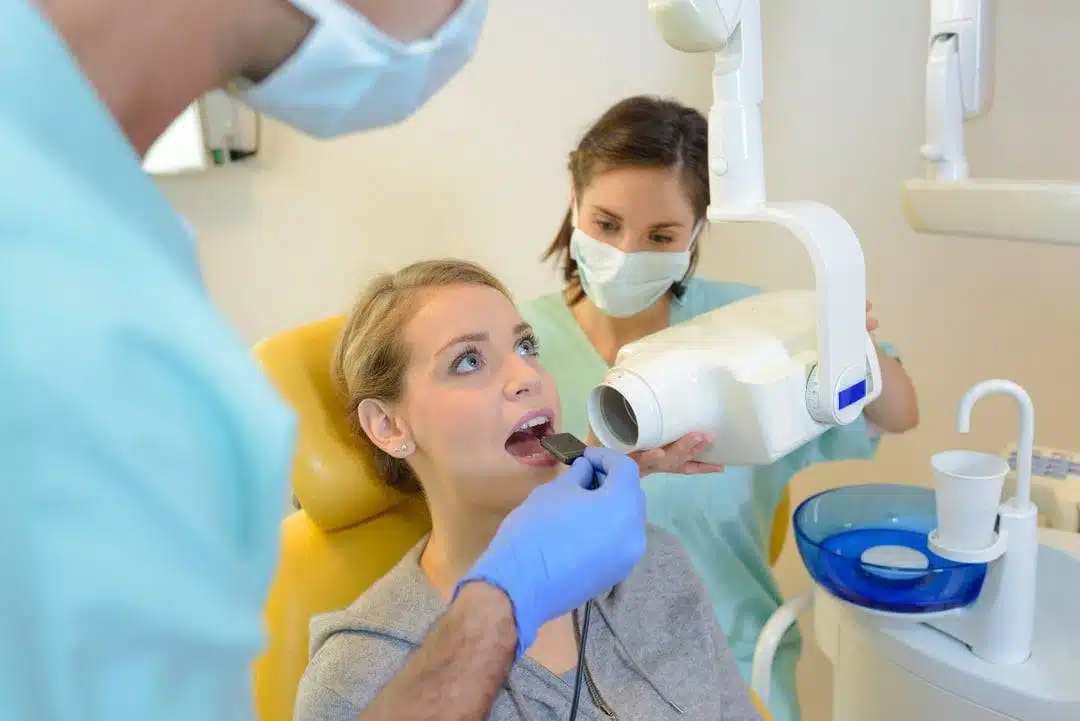 dental-x-rays-landing-page A woman sits in a dental chair while a dentist at a dental Grande Prairie clinic prepares her for a dental X-ray and an assistant positions the X-ray machine near her mouth.