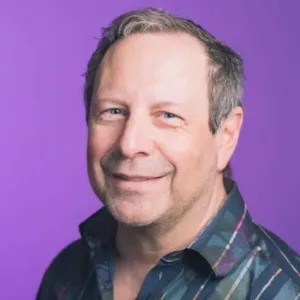A middle-aged man with short gray hair, wearing a plaid shirt, smiles at the camera against a plain purple background, showcasing his bright dental Grande Prairie smile.