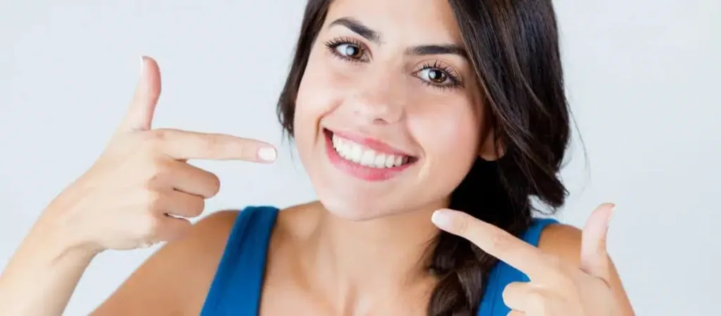 A woman with long dark hair smiles and points to her teeth with both index fingers, wearing a blue top against a plain background, highlighting her bright smile—perfect for dental Grande Prairie care.