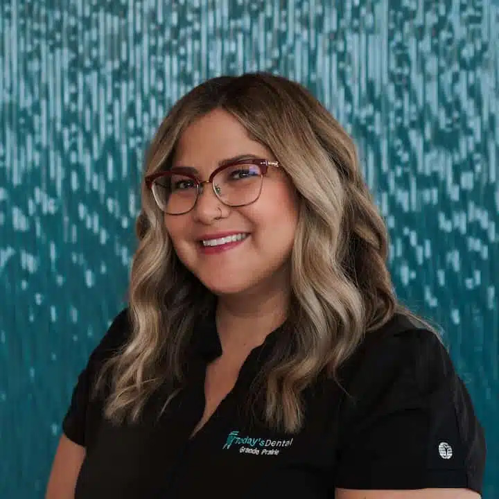 A woman with wavy blonde hair and glasses smiles while wearing a black dental grande prairie office shirt, standing in front of a blue patterned background.