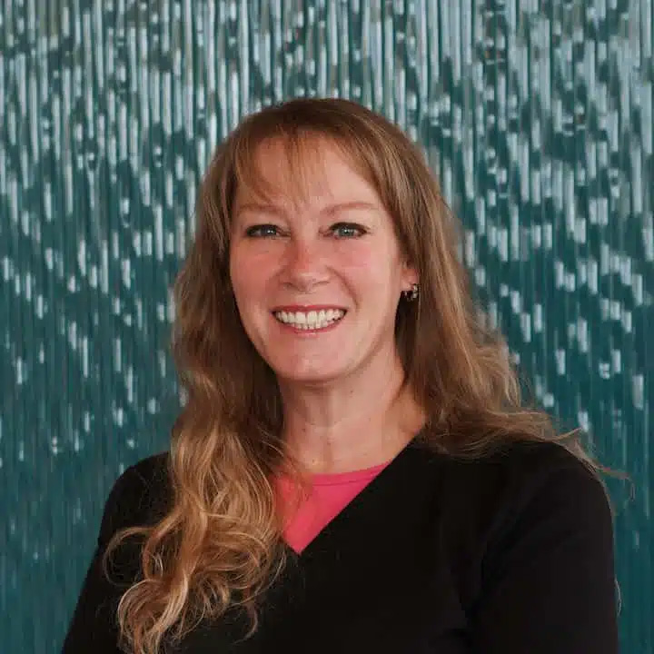 A woman with long, wavy blonde hair smiles at the camera. She is wearing a black top over a pink shirt, standing in front of a blue textured background, radiating confidence much like the team at dental Grande Prairie.