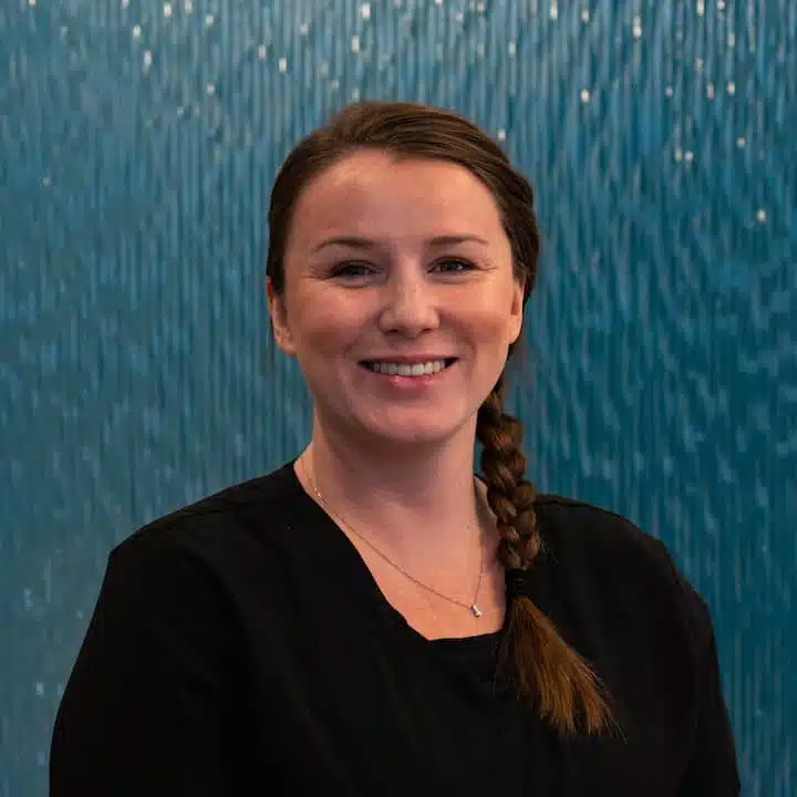 A woman with a brown braid, wearing a black top and necklace, smiles confidently in front of a blue textured background, reflecting the friendly atmosphere of dental Grande Prairie.