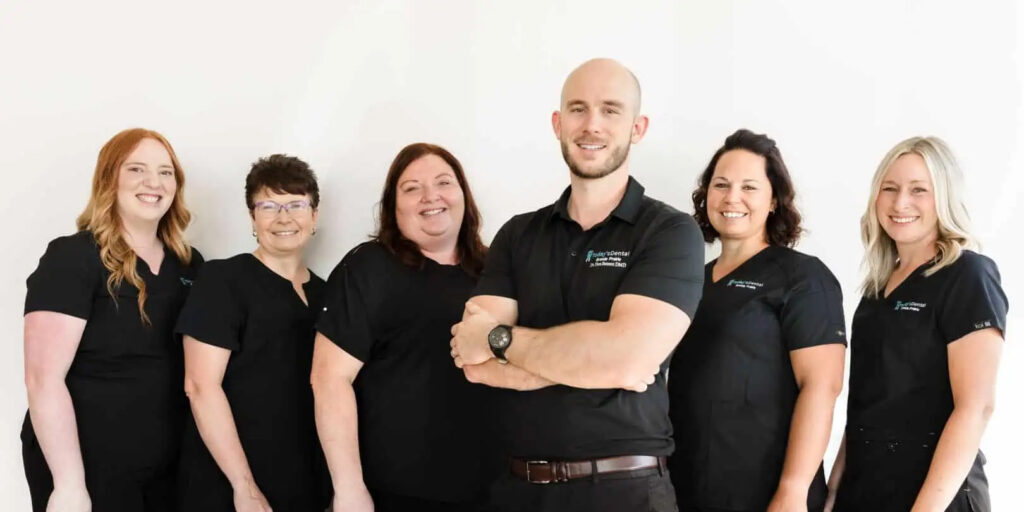 Six people in matching black uniforms stand in a row against a white background, smiling at the camera, representing the dental Grande Prairie team.