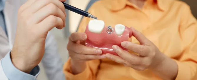 A dentist points at a dental implant model held by a patient, illustrating the placement of an artificial tooth in a gum replica.