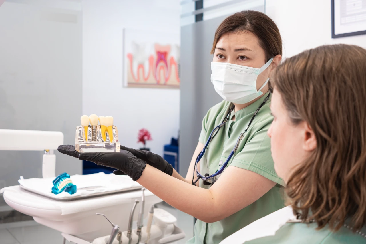 A dentist wearing a mask and gloves shows a dental model to a patient in a clinic. Dental tools and a tooth diagram are visible in the background.