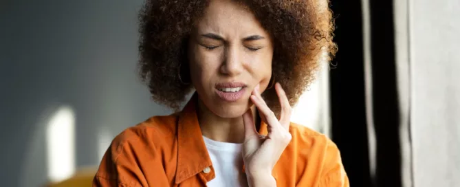 A woman with curly hair touches her cheek and closes her eyes, appearing to be in pain, possibly due to a toothache.