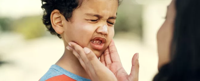 An adult applies sunscreen to a young boy's face. The boy’s eyes are closed and he appears uncomfortable.