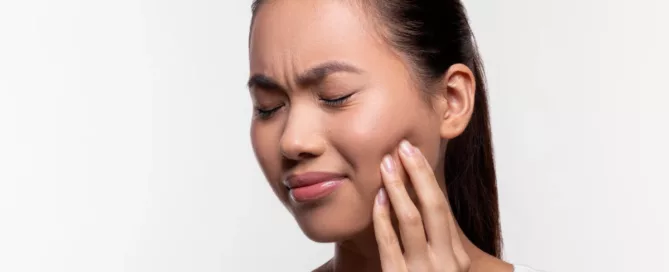 Woman with closed eyes touching her cheek, appearing to be in pain or discomfort, wearing a white tank top against a plain background.