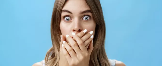 A woman with wide eyes covers her mouth with both hands, appearing surprised or shocked, against a solid light blue background.