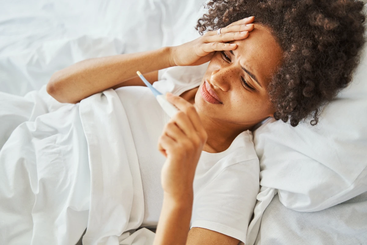 A person lying in bed holds a thermometer and touches their forehead, appearing unwell.
