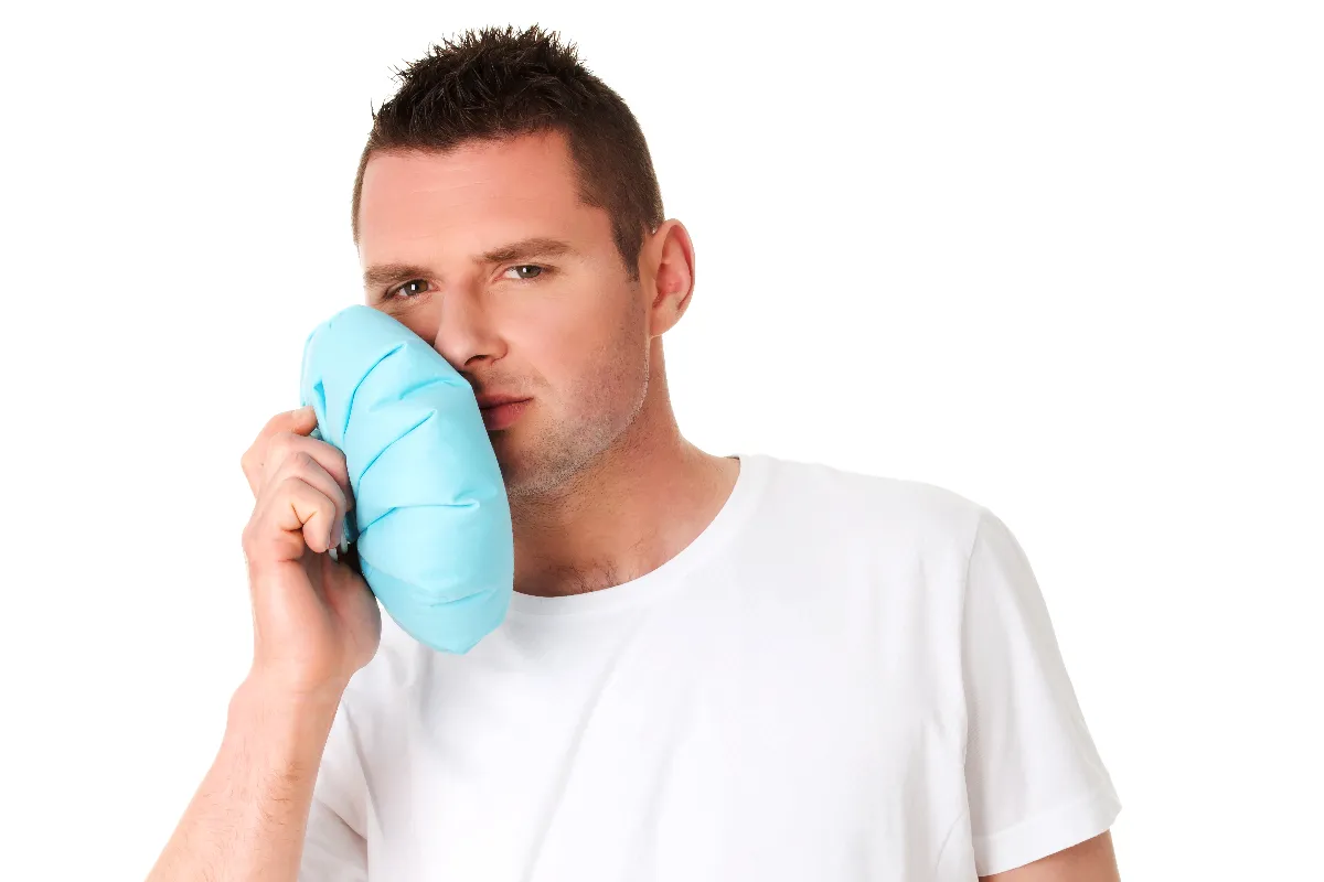 Man in a white t-shirt holding a blue ice pack to his cheek, appearing to treat facial pain, against a white background.