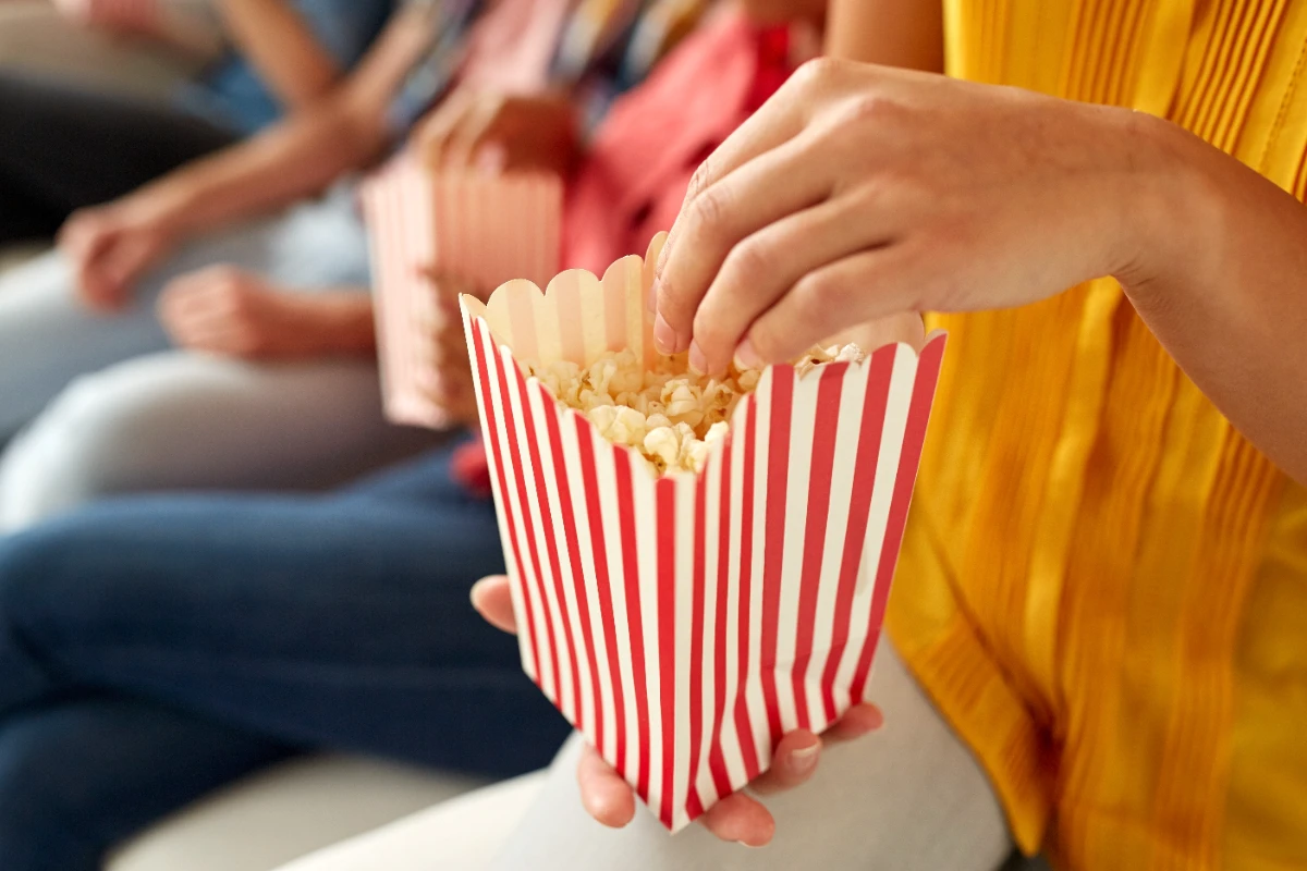 Person in a yellow top holds a red and white striped popcorn box and reaches in to grab some popcorn; other people are seated nearby.