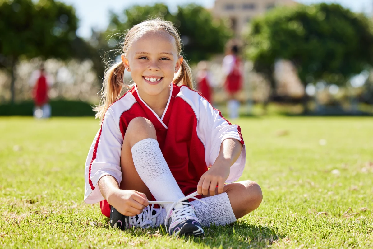 Smiling young girl in a red and white sports uniform sits on grass, tying her soccer cleats, with teammates visible in the blurred background.