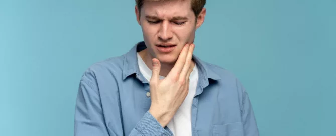 A young man in a blue shirt touches his jaw and appears to be in pain, standing against a plain blue background.