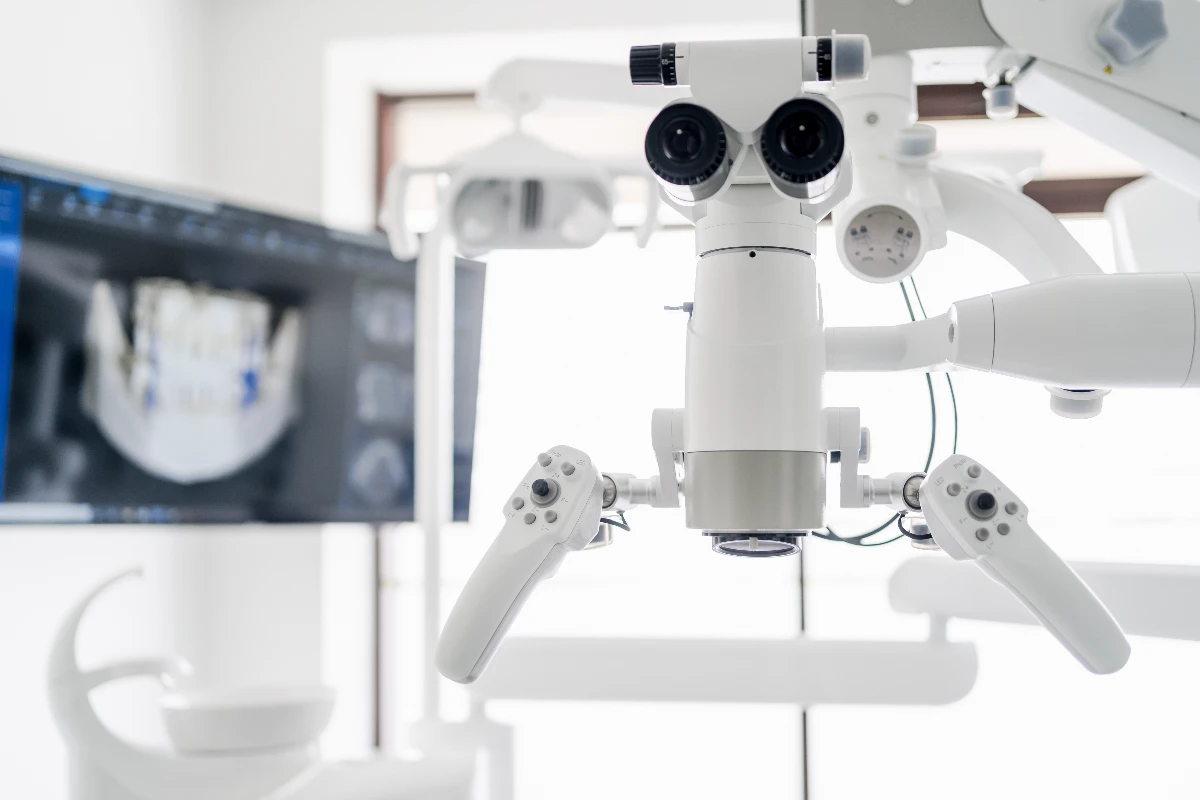 A close-up view of a dental microscope with dual eyepieces and control handles, positioned in front of a blurred computer screen showing dental imagery.