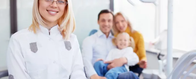 A female dentist stands in the foreground, smiling, with a family of three sitting together in the dental office background. Dental equipment is visible.